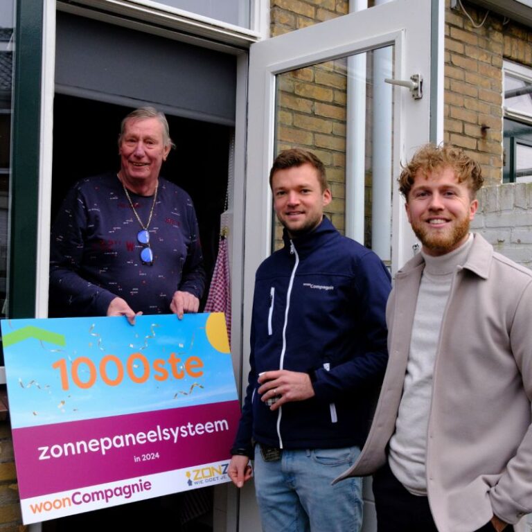 Foto van een medewerker van Zonzo, van de Wooncompagnie en van de bewoner van de woning die het 1000e zonnestroomsysteem dat jaar krijgt. De bewoner houd een bord vast.