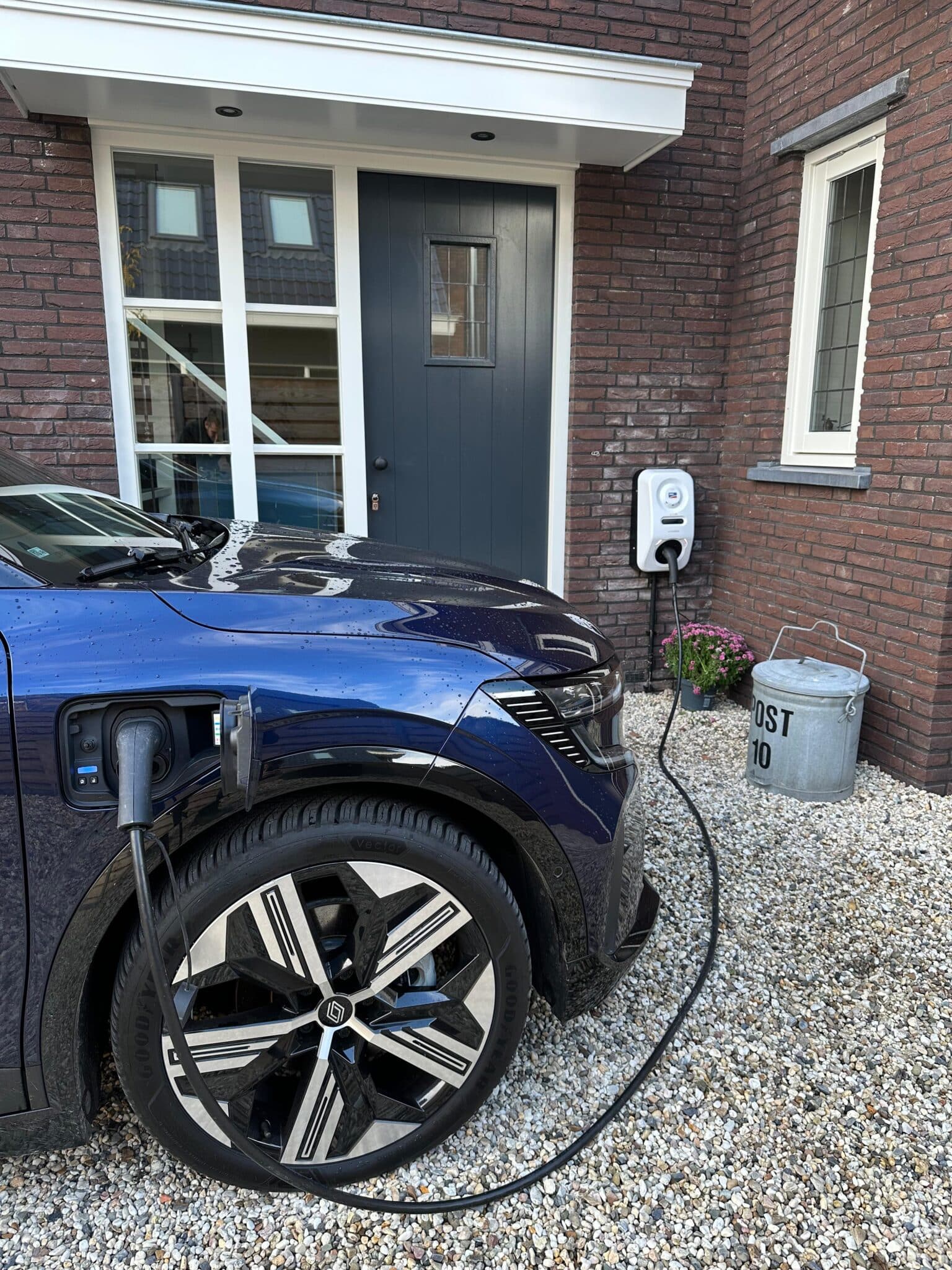 elektrische auto die wordt opgeladen met een lader die gebruikt maakt van zonnestroom. Deze lader met een extra lange kabel zorgt ervoor dat de auto kan worden opgeladen. Op de achtergrond hangt de lader tegen de muur van het huis.