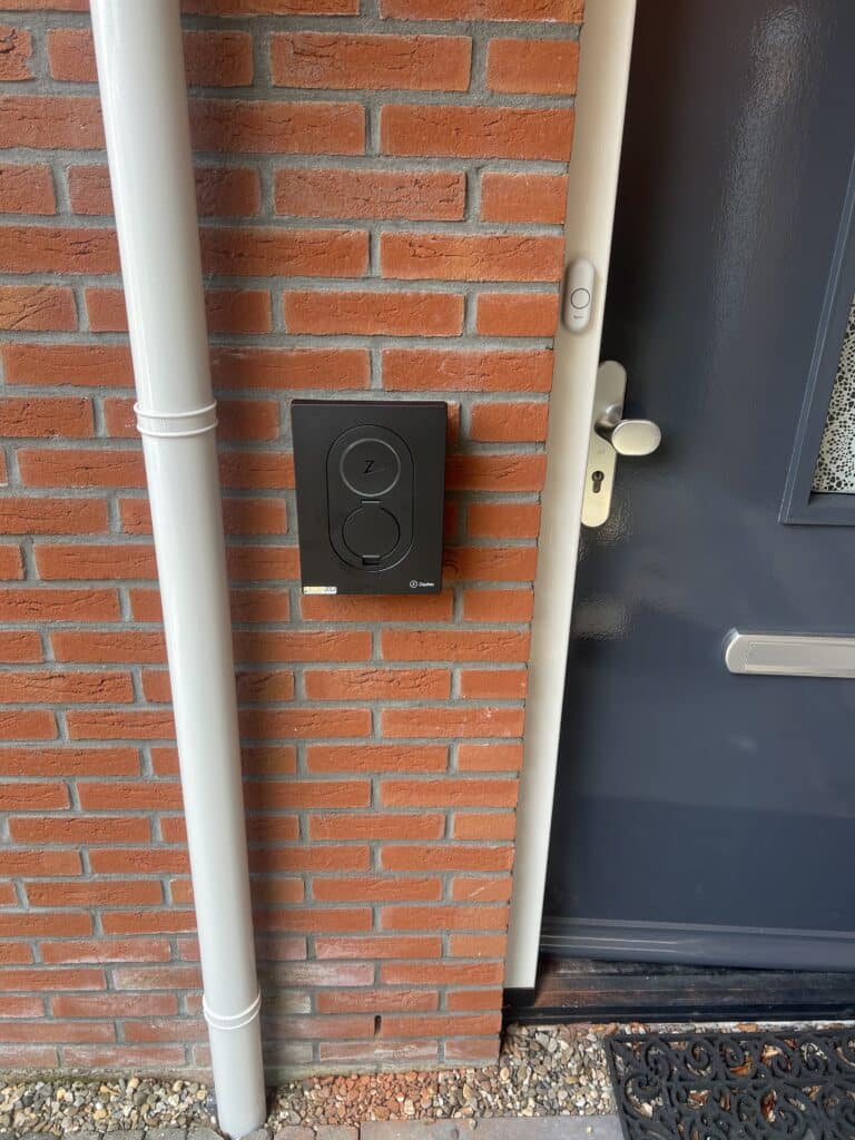 Foto van een elektrische laadpaal die bevestigt is aan een bakstenen muur naast de voordeur.